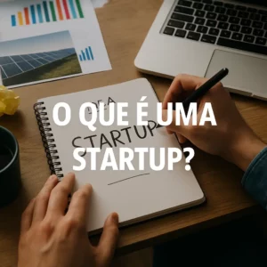 Pessoa a escrever "startup" num caderno sobre uma secretária com computador portátil, gráficos e caneca, com o texto “O QUE É UMA STARTUP?” ao centro da imagem.